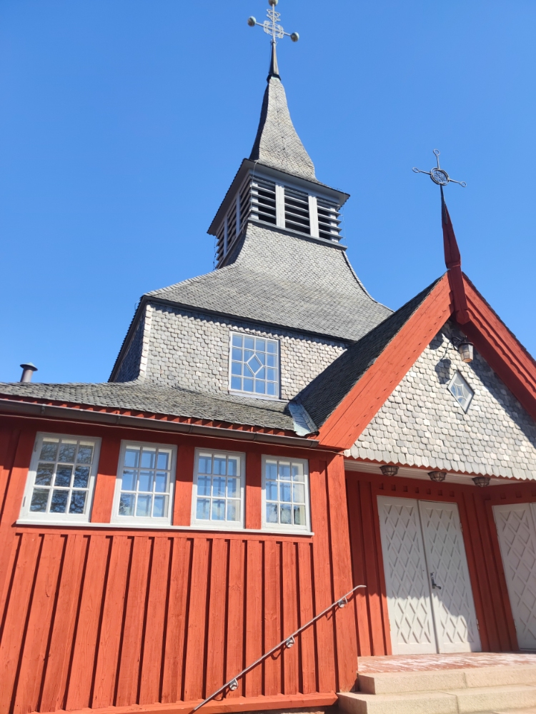 Röd träkyrka med ett brant, grått spåntak och en hög spira mot en klarblå himmel.