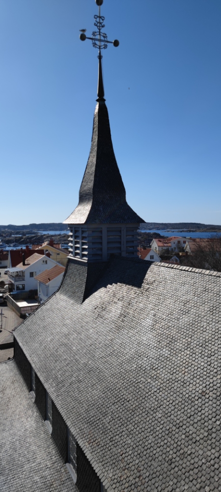 Flygfoto över en kyrka med ett brant skiffertak och högt torn, mot en klarblå himmel och en kuststad.