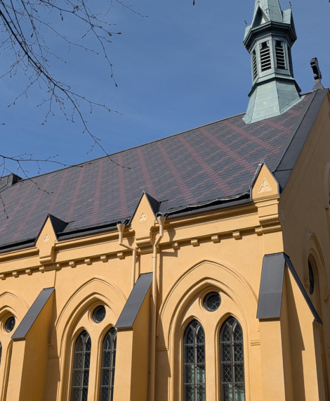 En gul byggnad med spetsiga välvda fönster, dekorativa lister och ett brant, spånbeklätt tak med en hög, smal spira. Grenar från ett närliggande träd syns i bildens övre vänstra hörn.